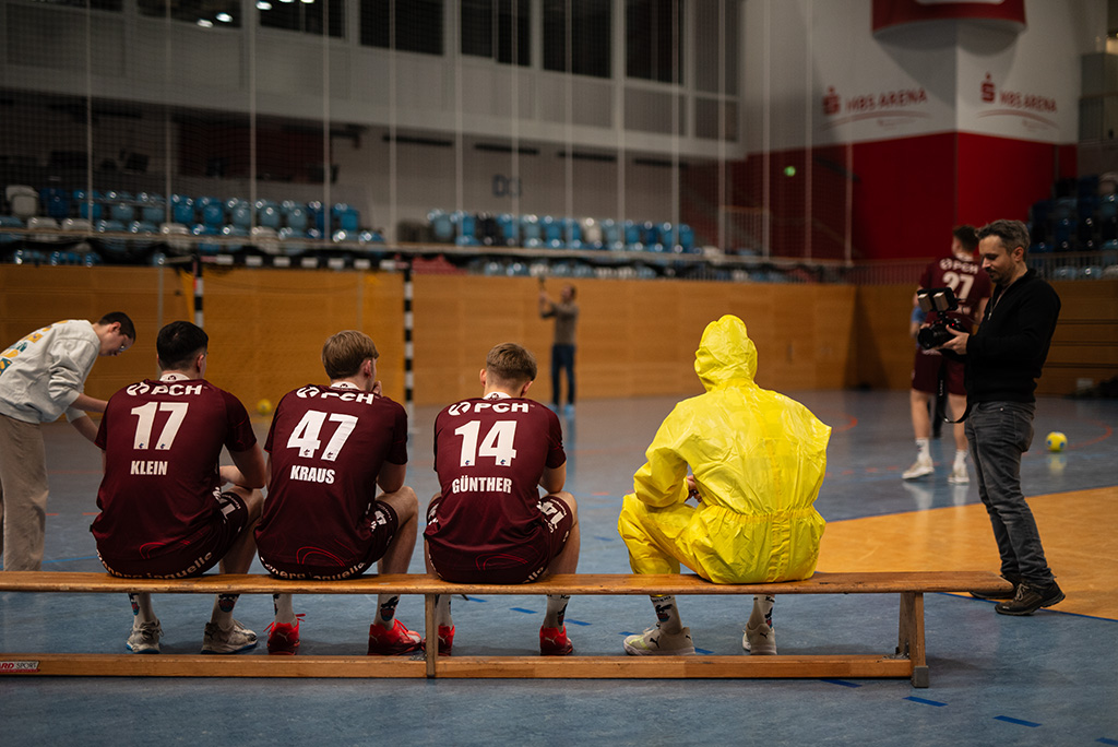 4 Spieler vom VfL Potsdam sitzen in einer Sporthalle auf einer Bank, 3 im Trikot, einer hat einen gelben Anzug an. Auf dem Feld stehen 3 Menschen mit Kameras verteilt.